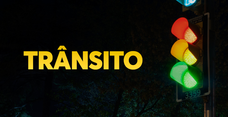 Imagem com fundo escuro e noturno contendo o texto TRÂNSITO em letras maiúsculas na cor amarela, posicionado no lado esquerdo. No lado direito, sobressai-se um semáforo aceso em um poste, exibindo as luzes vermelha, amarela e verde contra o fundo escuro de árvores e da noite.