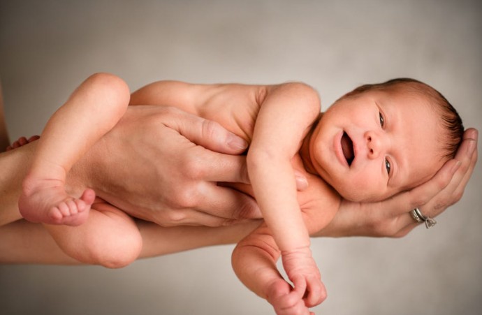 Fotografia em plano médio com iluminação suave, mostrando um bebê recém-nascido e sem roupas sendo segurado horizontalmente pelas mãos de um adulto. O bebê está deitado de costas, com a cabeça apoiada na mão direita do adulto e as costas e pernas amparadas pela mão esquerda. Ele tem cabelos escuros, olhos entreabertos e a boca levemente aberta. O fundo é neutro e desfocado em tons de bege.